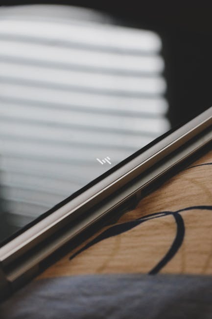 A close-up of a silver HP laptop resting on a wooden surface, with the lid slightly open revealing the keyboard. In the background, a window with partially closed white blinds diffuses natural light i
